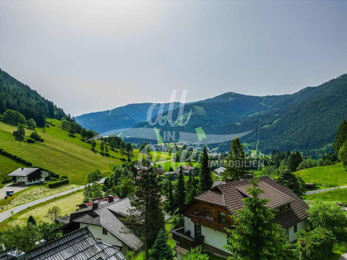 Weitläufiger Blick auf ein Dorf in einem grünen Tal, umgeben von bewaldeten Berghängen.
