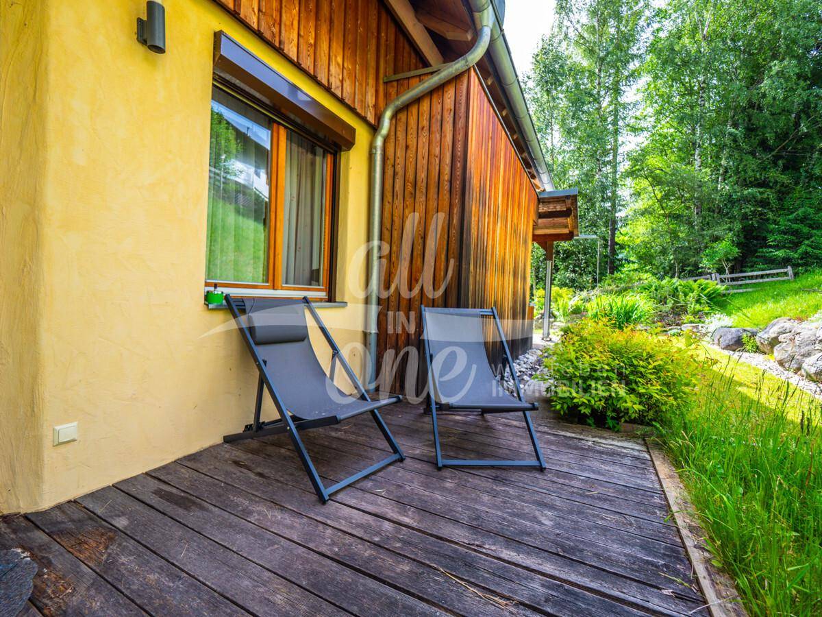 Holzterrasse mit zwei Liegestühlen, umgeben von grüner Natur und der Hausfassade.