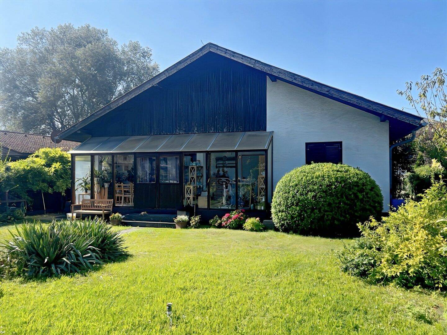 Hausfassade mit Wintergarten und weitläufigem Gartenbereich unter blauem Himmel.