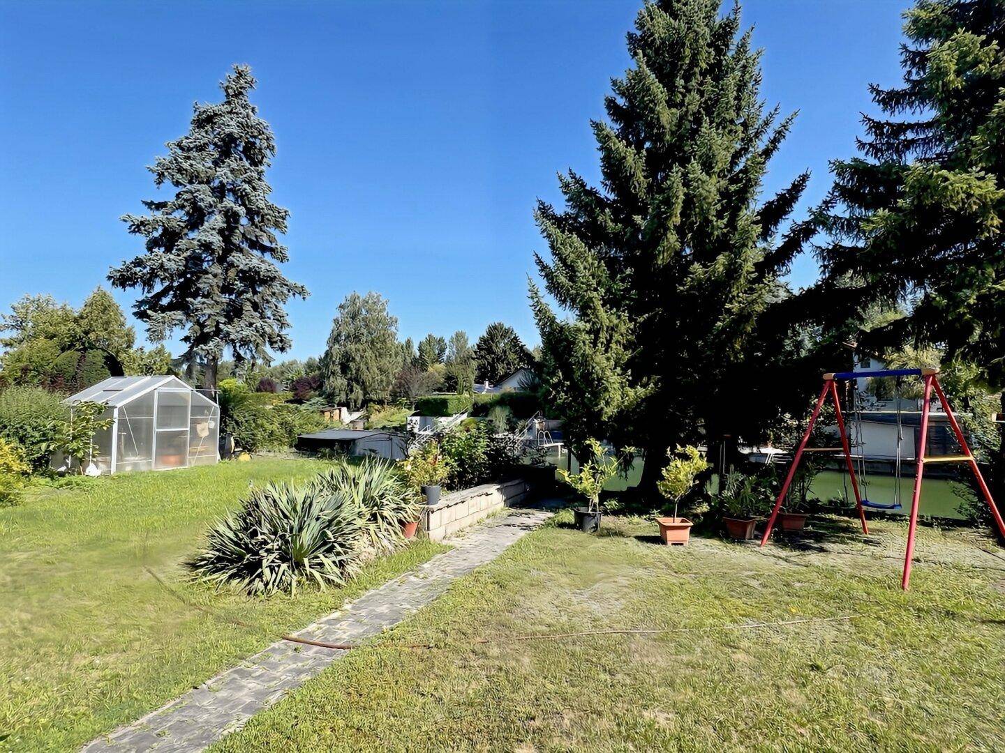 Weitläufiger Garten mit Rasenfläche, Schaukel und Gewächshaus unter strahlend blauem Himmel.