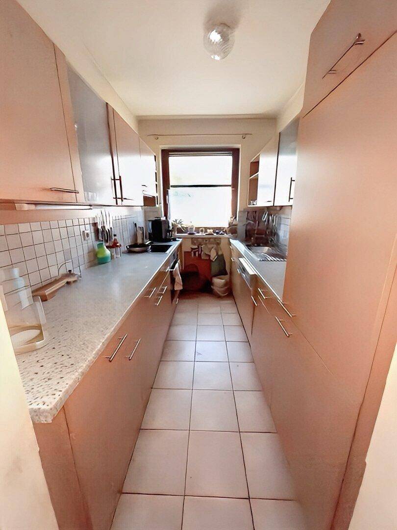 Schmale Küche mit beigen Schränken, Fliesenboden und Fenster für Tageslicht.