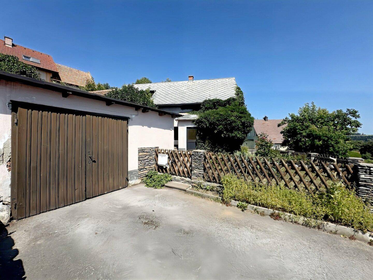 Garage mit Holztor und Zufahrt, angrenzend an das Wohnhaus mit rustikalem Zaun.