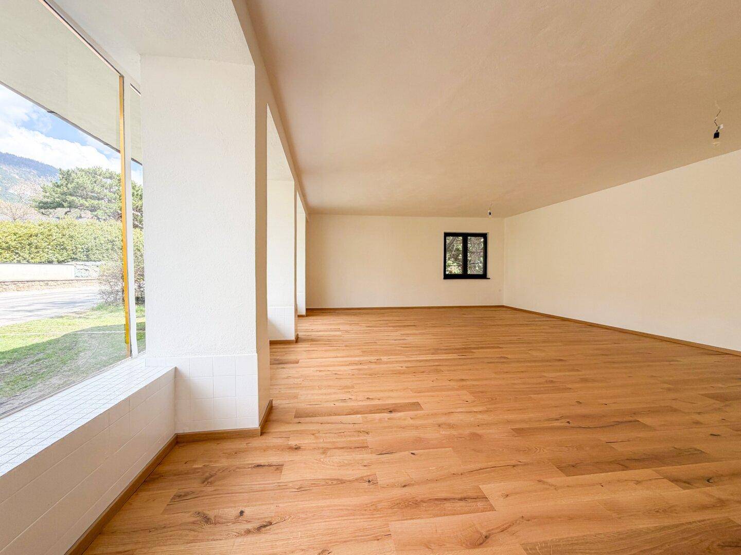 Großer, heller Raum mit Holzboden und breiten Fenstern, die viel Tageslicht hereinlassen.