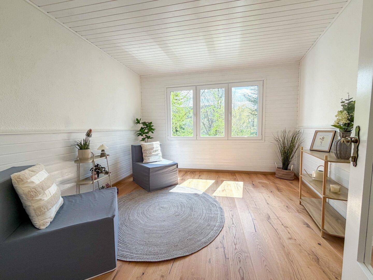 Helles Wohnzimmer mit Holzboden, weiß getäfelten Wänden und Decke sowie Blick ins Grüne.