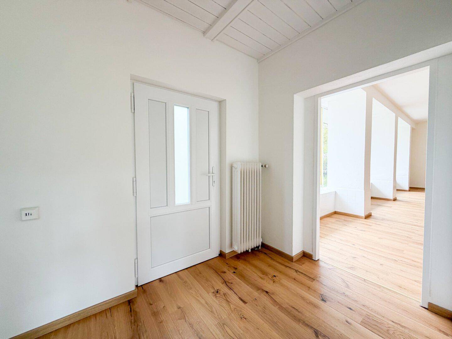 Heller Flur mit Holzboden und weißer Tür, der zu weiteren Räumen mit ähnlichem Boden führt.