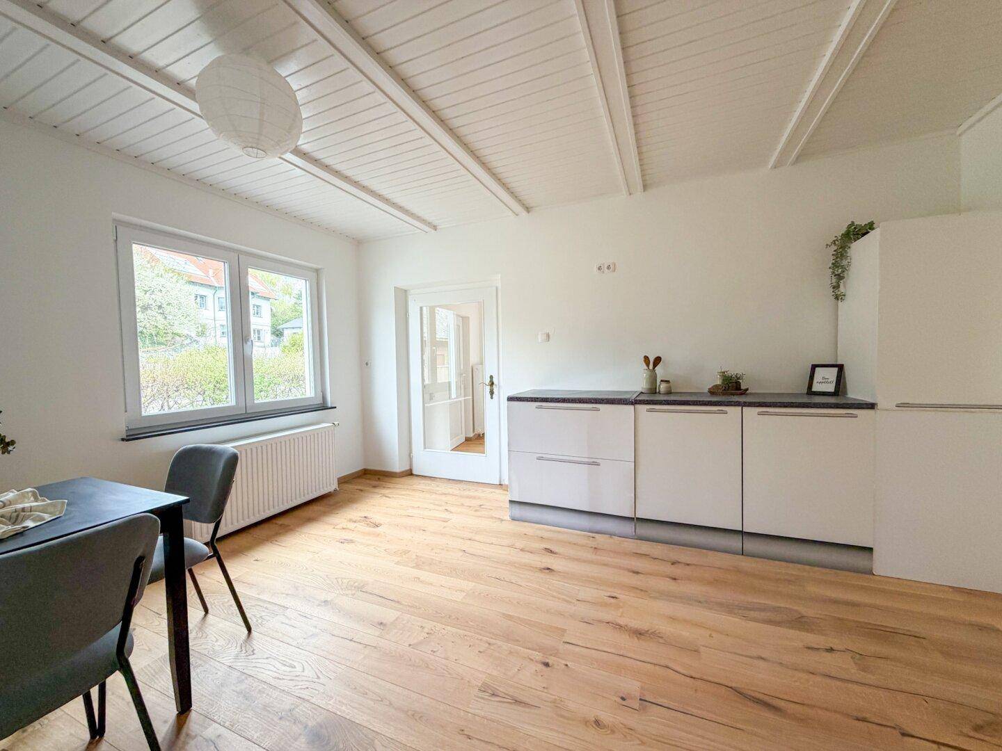 Essbereich mit Küchenzeile, Holzboden und Fenster, das viel Tageslicht hereinlässt.