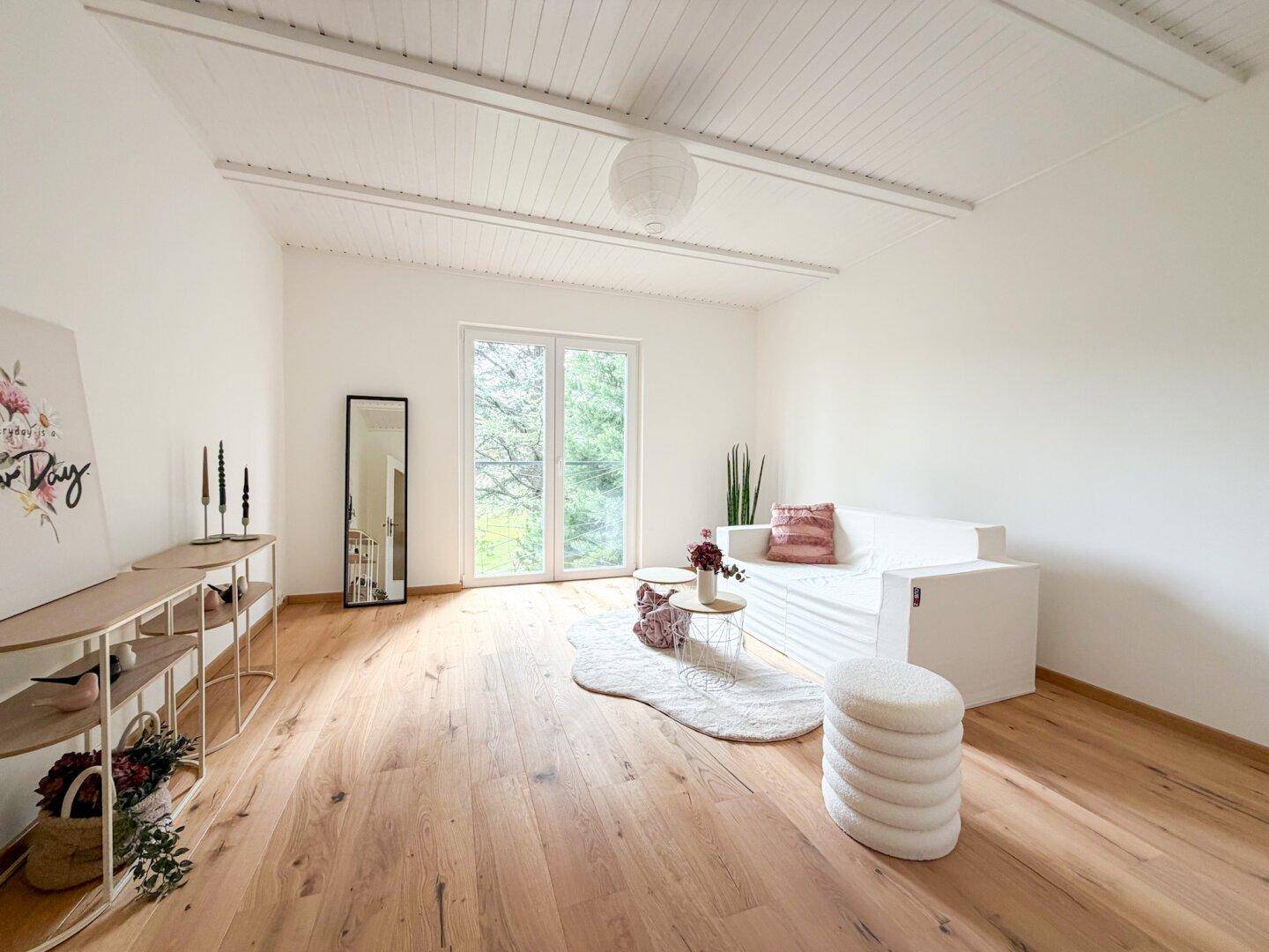 Lichtdurchflutetes Wohnzimmer mit Holzboden, weißer Decke und Zugang zu einem Balkon oder Garten.