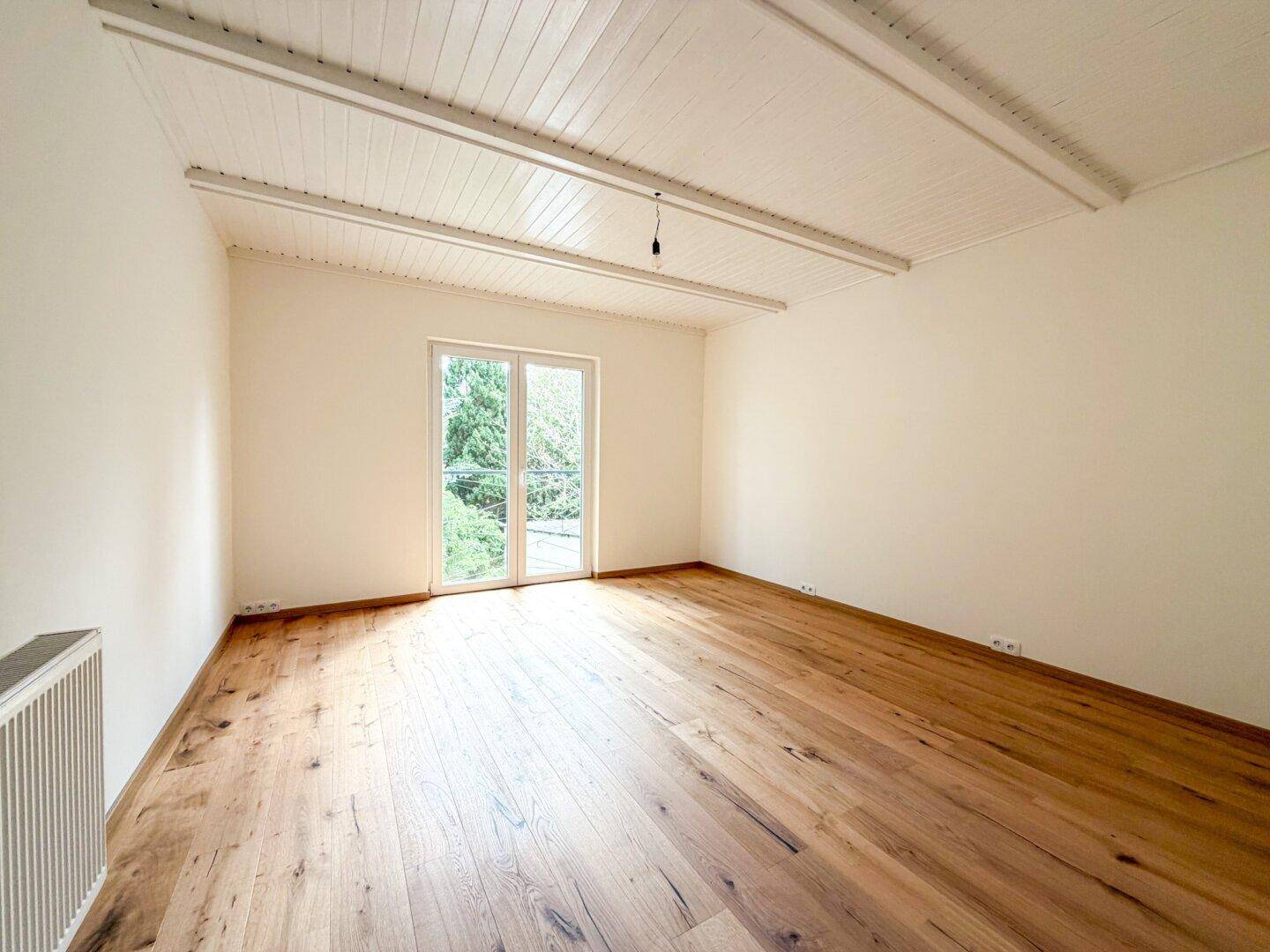 Leeres Schlafzimmer mit Holzboden, weiß gestrichenen Wänden und Zugang zu einem Balkon oder Garten.
