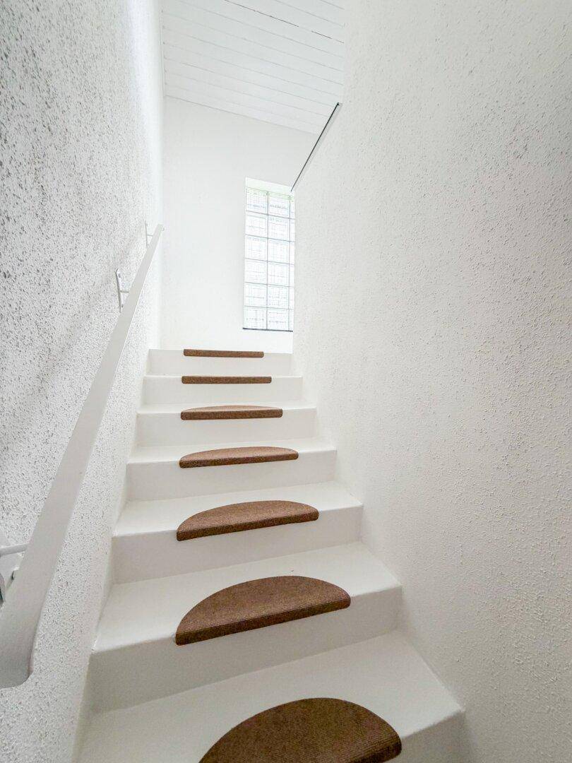 Weiß gestrichene Treppe mit Teppichstufen und einem Fenster, das Tageslicht in den Treppenbereich bringt.