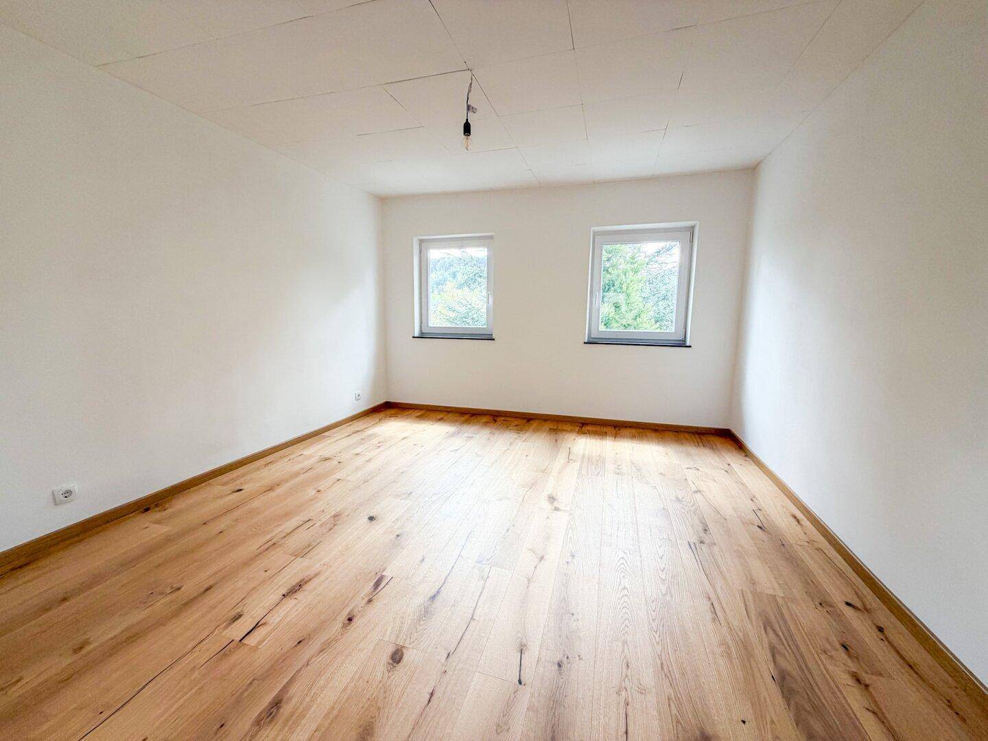 Leeres Schlafzimmer mit Holzboden und zwei Fenstern, die einen Blick ins Grüne bieten.