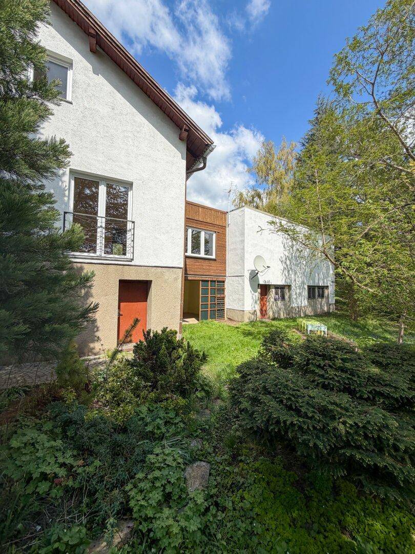 Rückansicht des Hauses mit weißer Fassade und Holzelementen, umgeben von üppigem Grün und Bäumen.