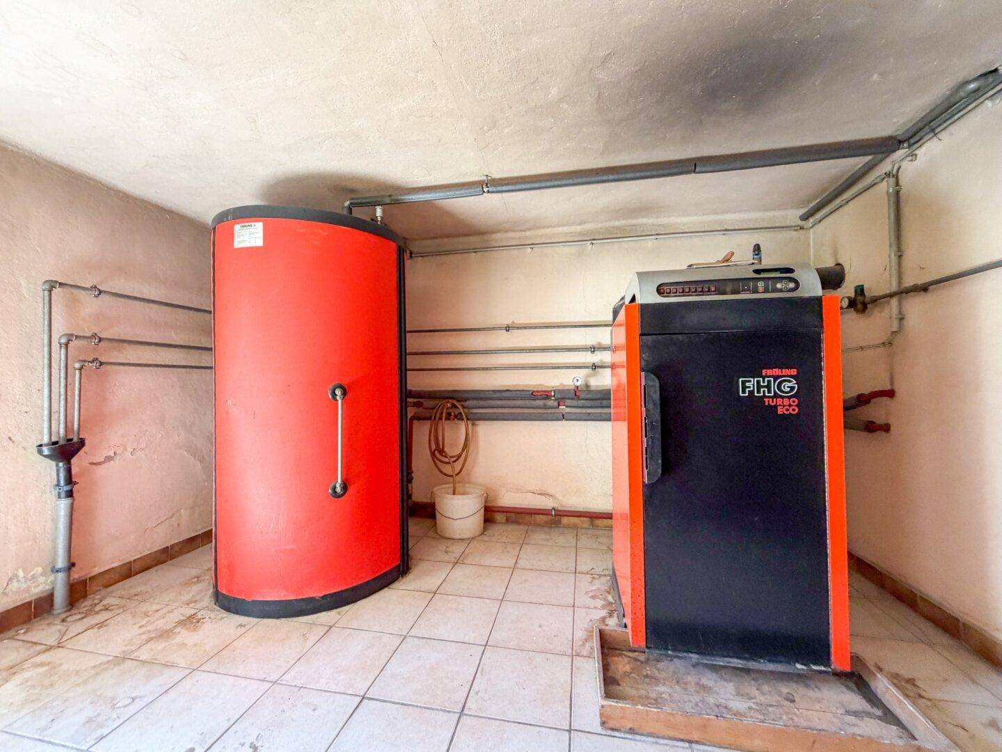 Heizungsraum mit großem Warmwasserspeicher und Heizkessel, umgeben von Rohrleitungen.