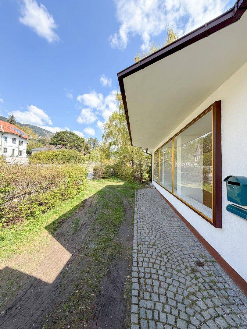 Seitenansicht des Hauses mit großen Fenstern und gepflastertem Weg, umgeben von grüner Landschaft.