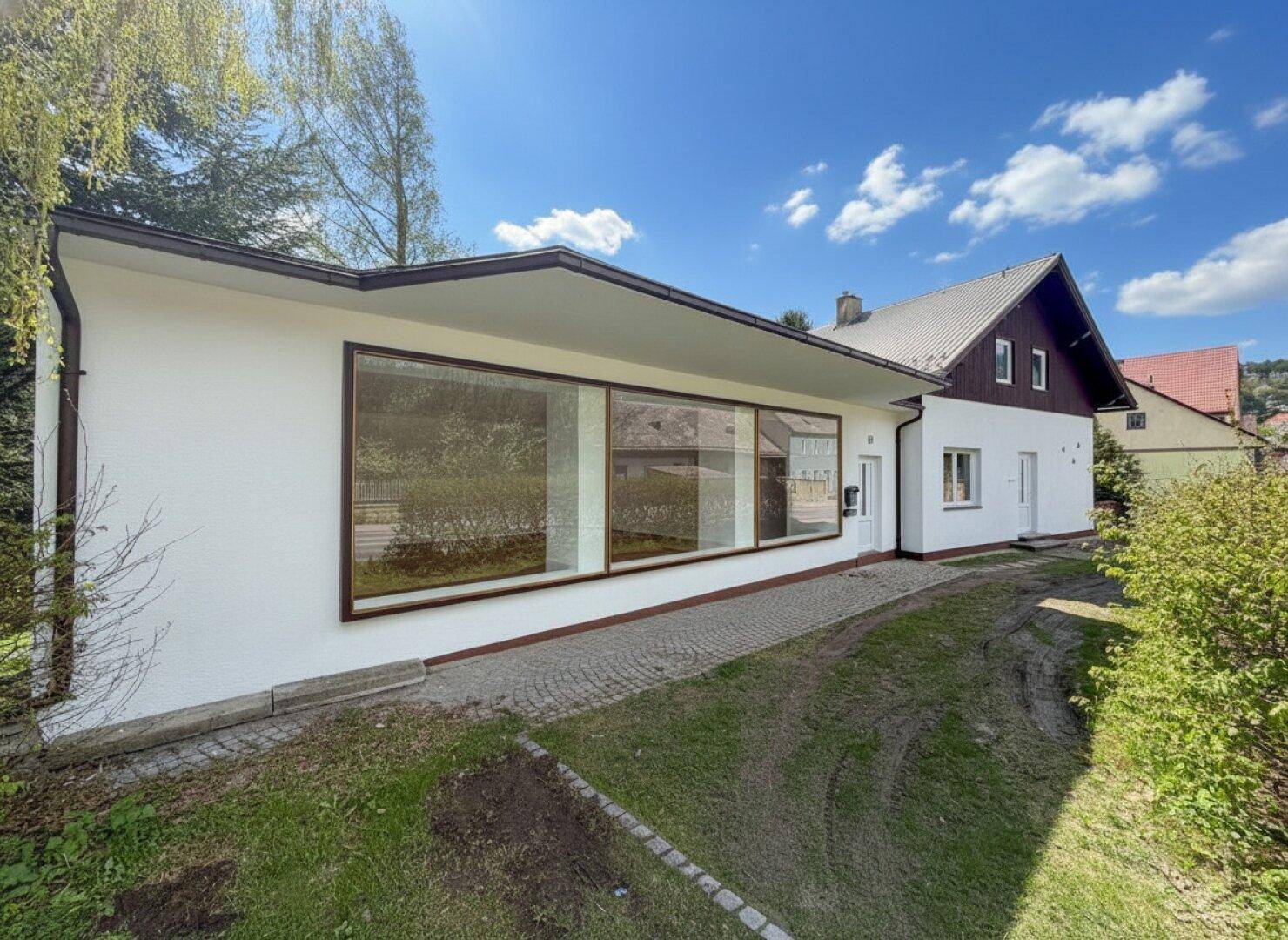 Seitenansicht des Hauses mit großen Fenstern und weißer Fassade, umgeben von viel Grün und einem gepflasterten Weg.