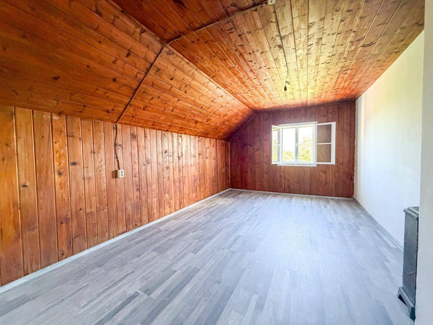 Dachzimmer mit Holzwänden und -decke, Laminatboden und einem Fenster, das Tageslicht hereinlässt.