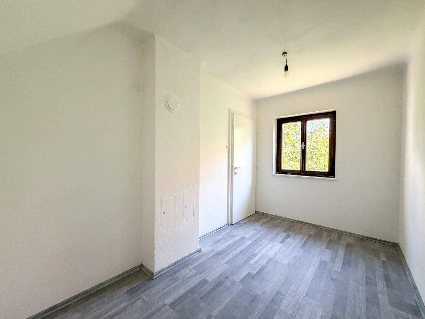 Leeres Zimmer mit Laminatboden und einem Fenster, das viel natürliches Licht hereinlässt.