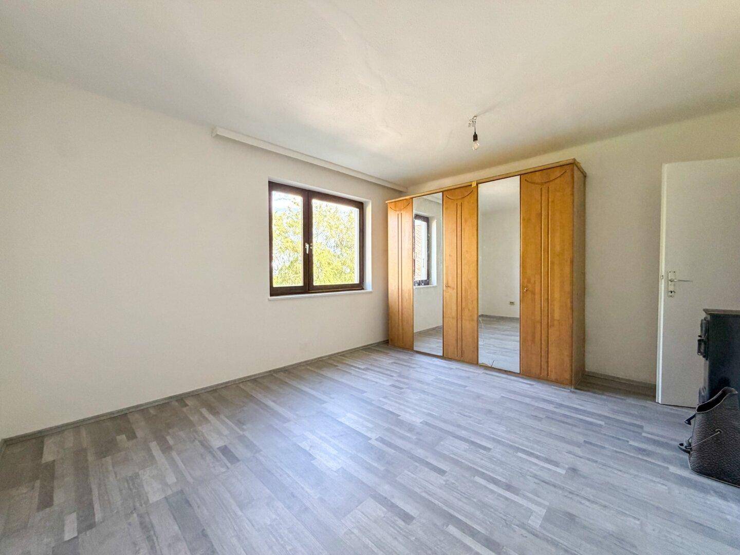 Schlafzimmer mit Laminatboden, einem großen Fenster und einem Spiegelschrank an der Wand.