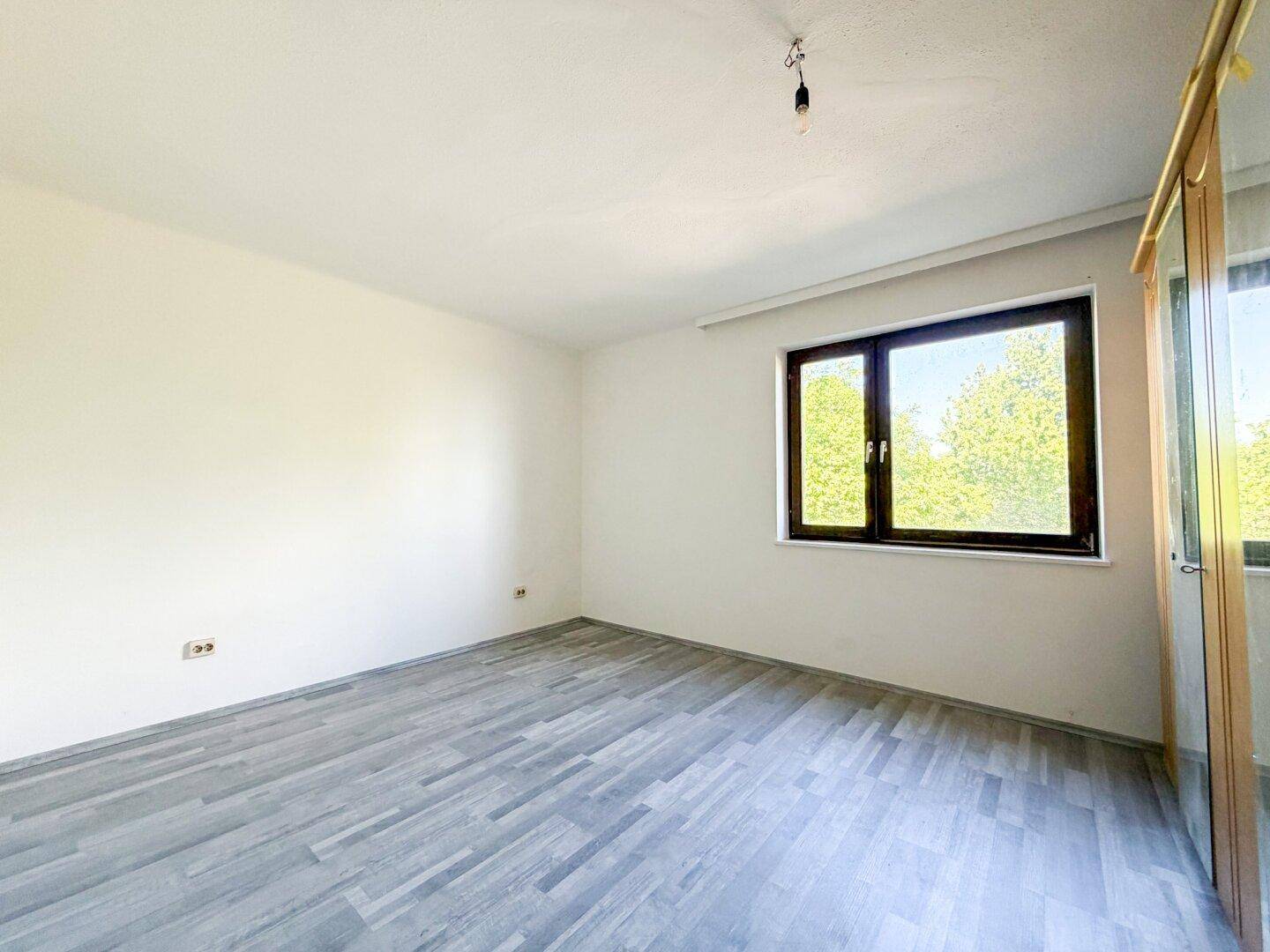 Helles Schlafzimmer mit Laminatboden und einem großen Fenster, das viel Tageslicht hereinlässt.