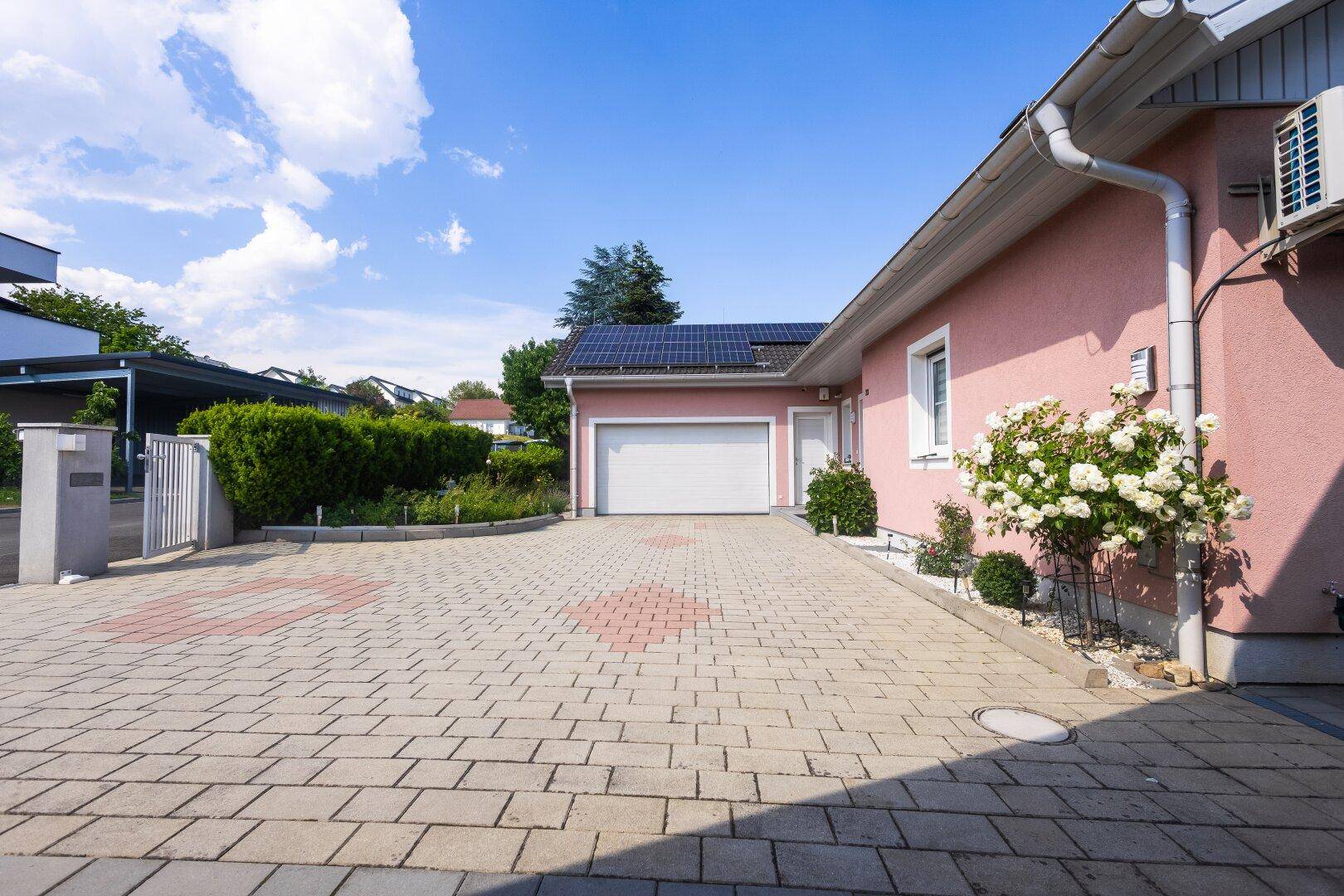 Vorderansicht des Hauses mit gepflasterter Einfahrt, Garage und blühenden Sträuchern.