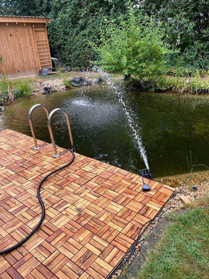 Natürlicher Schwimmteich mit Holzdeck, Einstiegsleiter und Wasserspiel in grüner Umgebung.