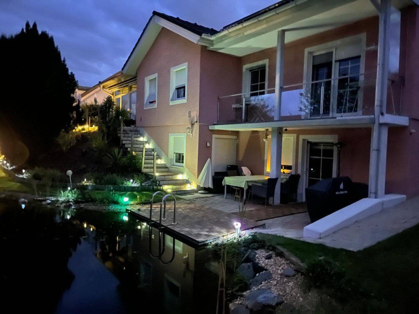 Beleuchtetes Haus mit Terrasse, Balkonen und naturnahem Teich im Garten bei Dämmerung.