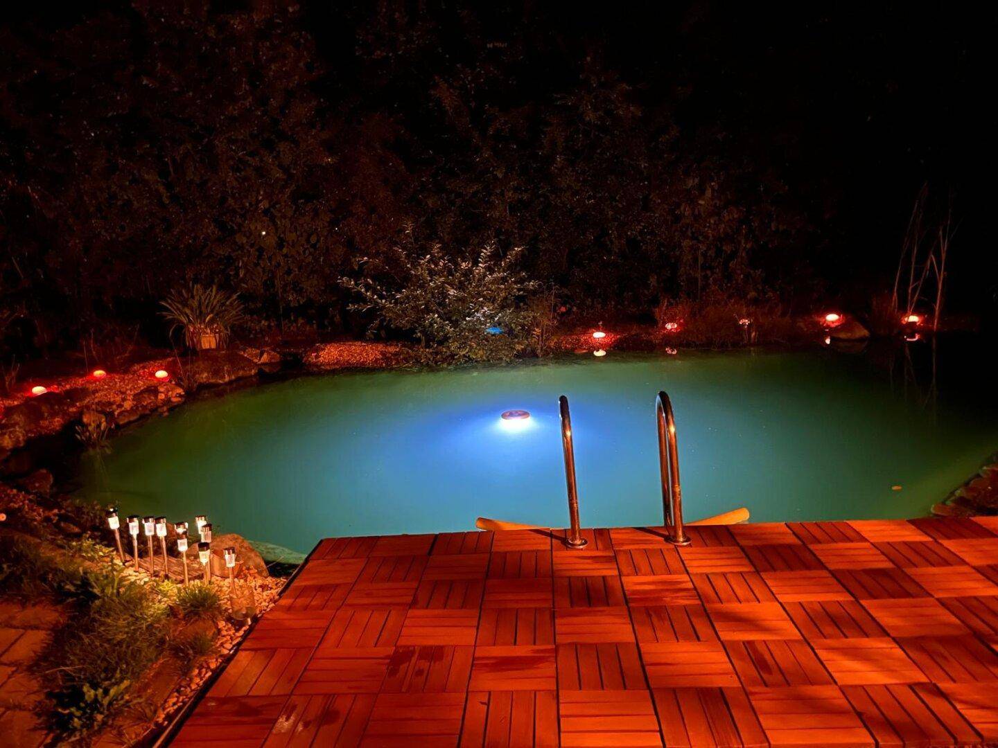 Stimmungsvoll beleuchteter Teichbereich mit Holzdeck und dekorativen Lichtern bei Nacht.
