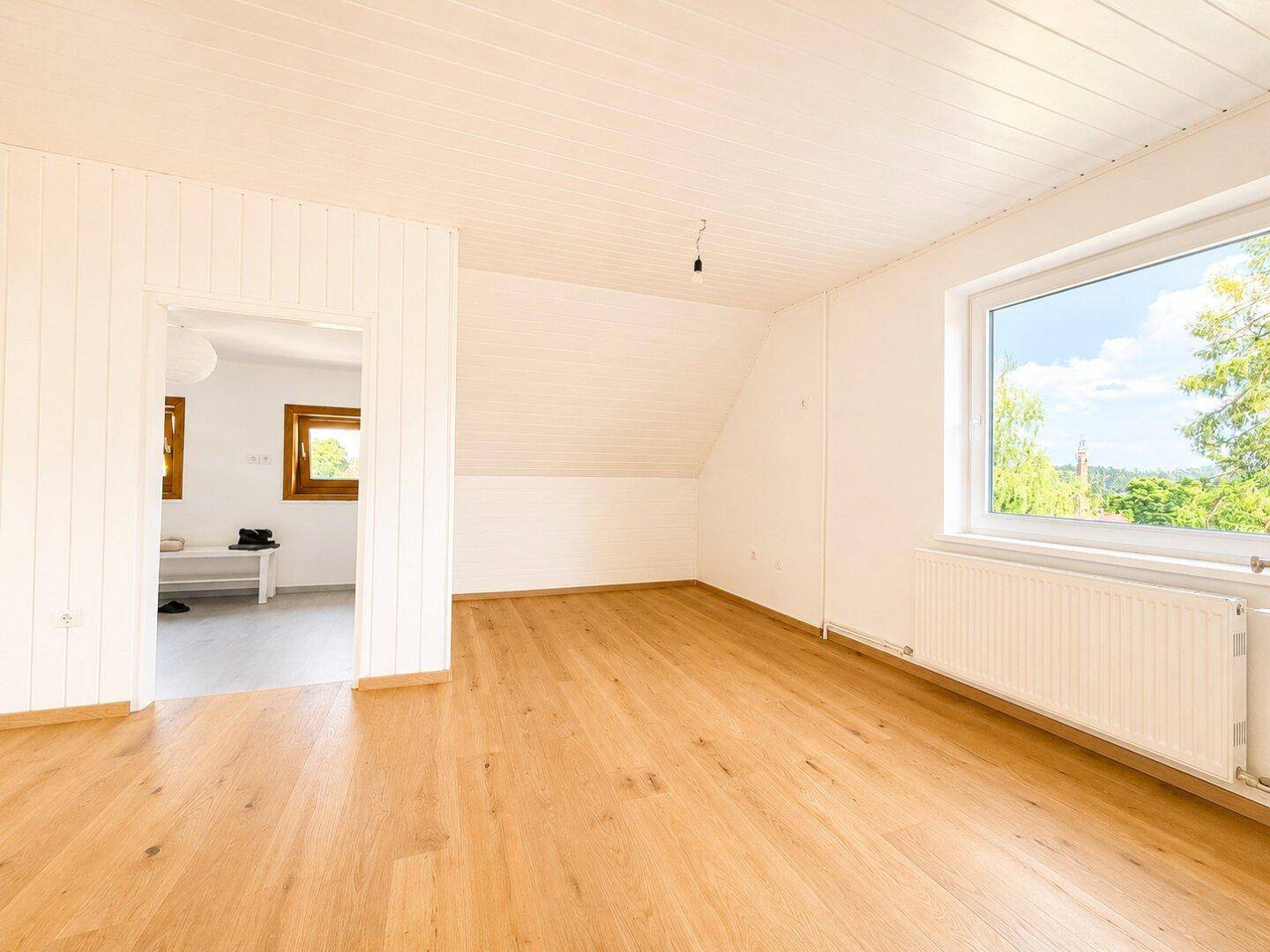 Helles Zimmer im Dachgeschoss mit Holzboden, weißer Holzdecke und großem Fenster.