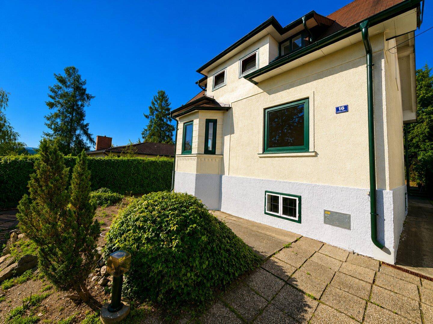 Seitenfassade des Hauses mit Erkerfenster und grünen Akzenten, umgeben von gepflegter Bepflanzung.