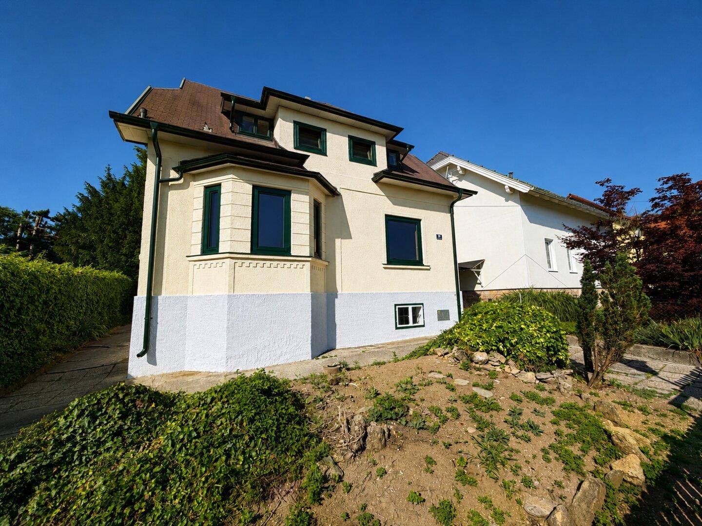 Hausfassade mit markantem Erker und umliegendem Gartenbereich unter blauem Himmel.