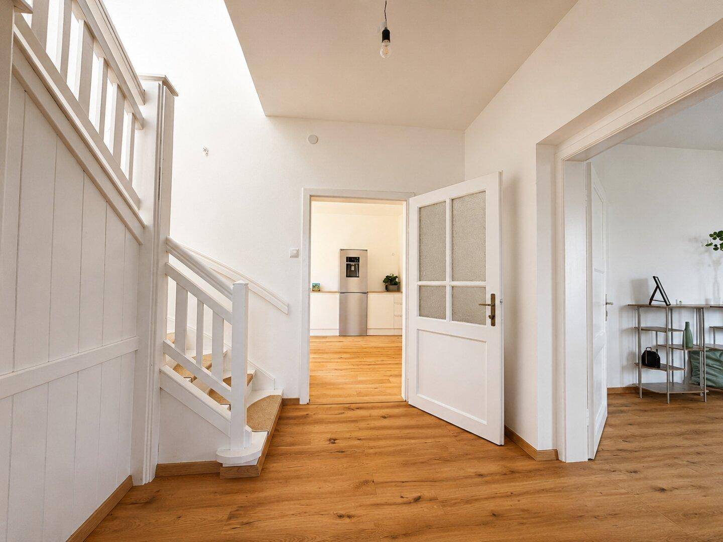 Heller Flur mit Holzboden, weißer Treppe und offener Verbindung zur Küche und Wohnbereich.