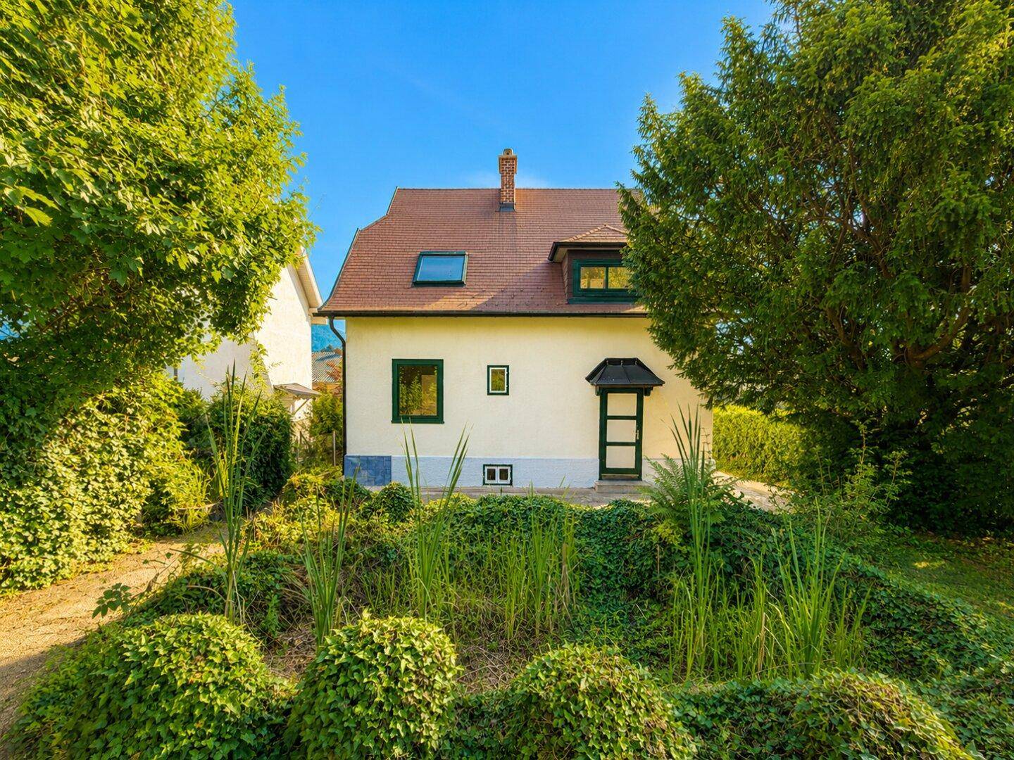 Vorderansicht des Hauses mit grünen Fensterrahmen und einem sonnigen, grünen Vorgarten.