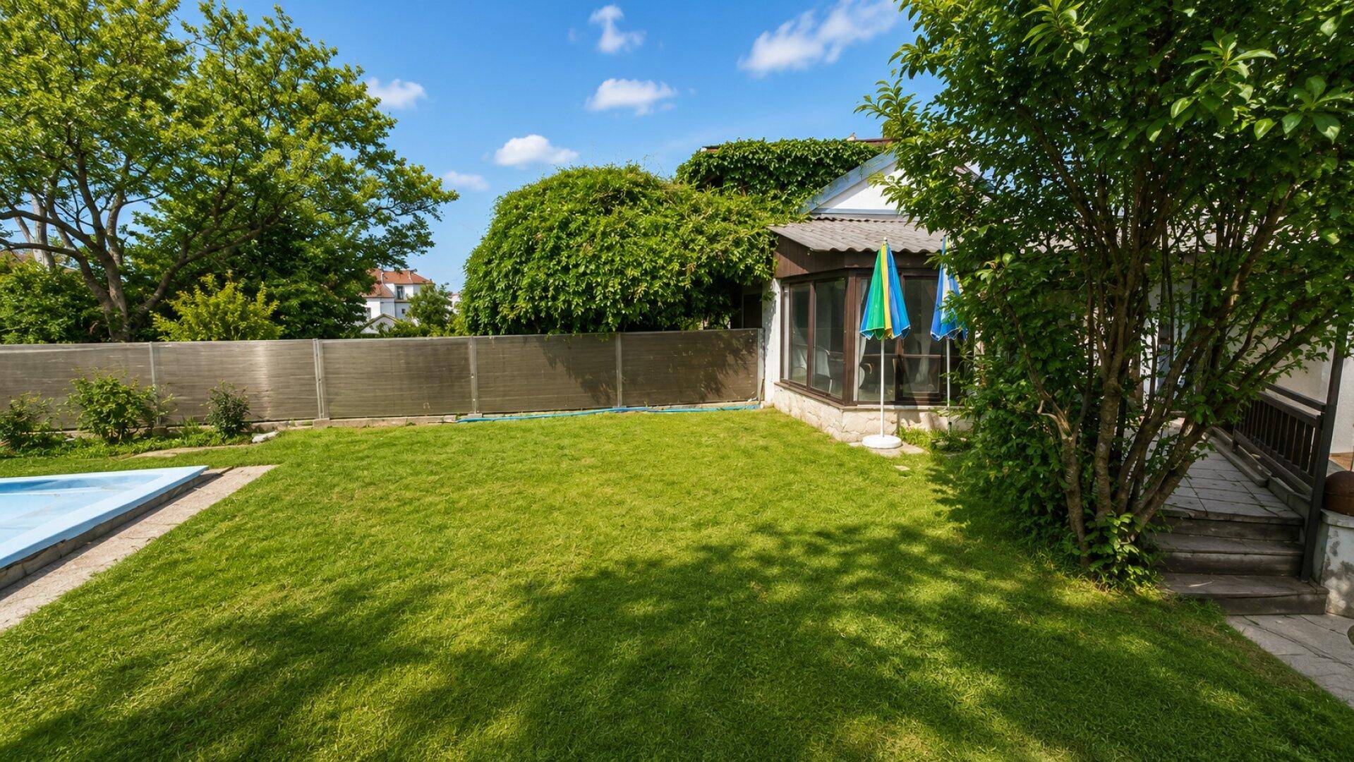 Grüner Gartenbereich mit kleinem Pool, Rasenfläche und einem Wintergarten am Haus unter blauem Himmel.