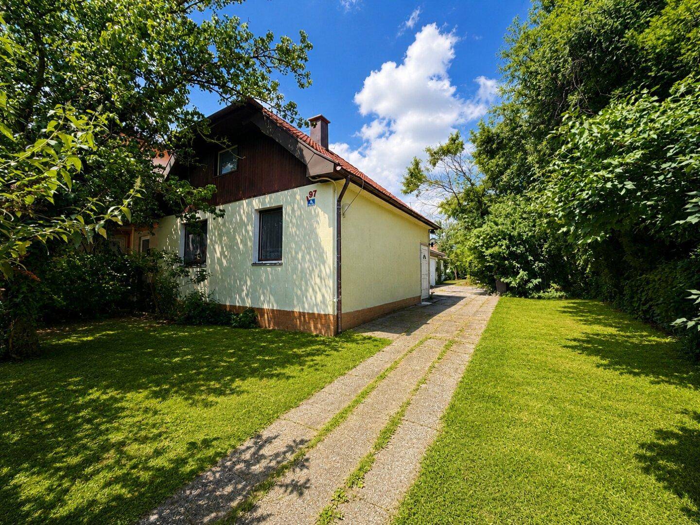 Hausfassade mit hellem Anstrich, umgeben von einem grünen Garten und einer gepflasterten Auffahrt unter blauem Himmel.
