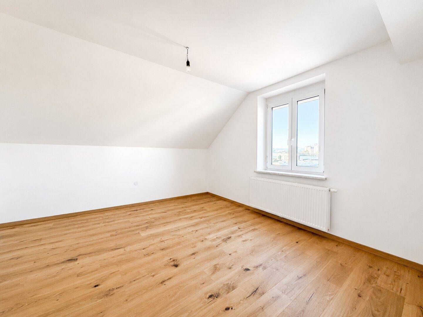 Helles Schlafzimmer im Dachgeschoss mit Holzboden, weißen Wänden und Fenster.