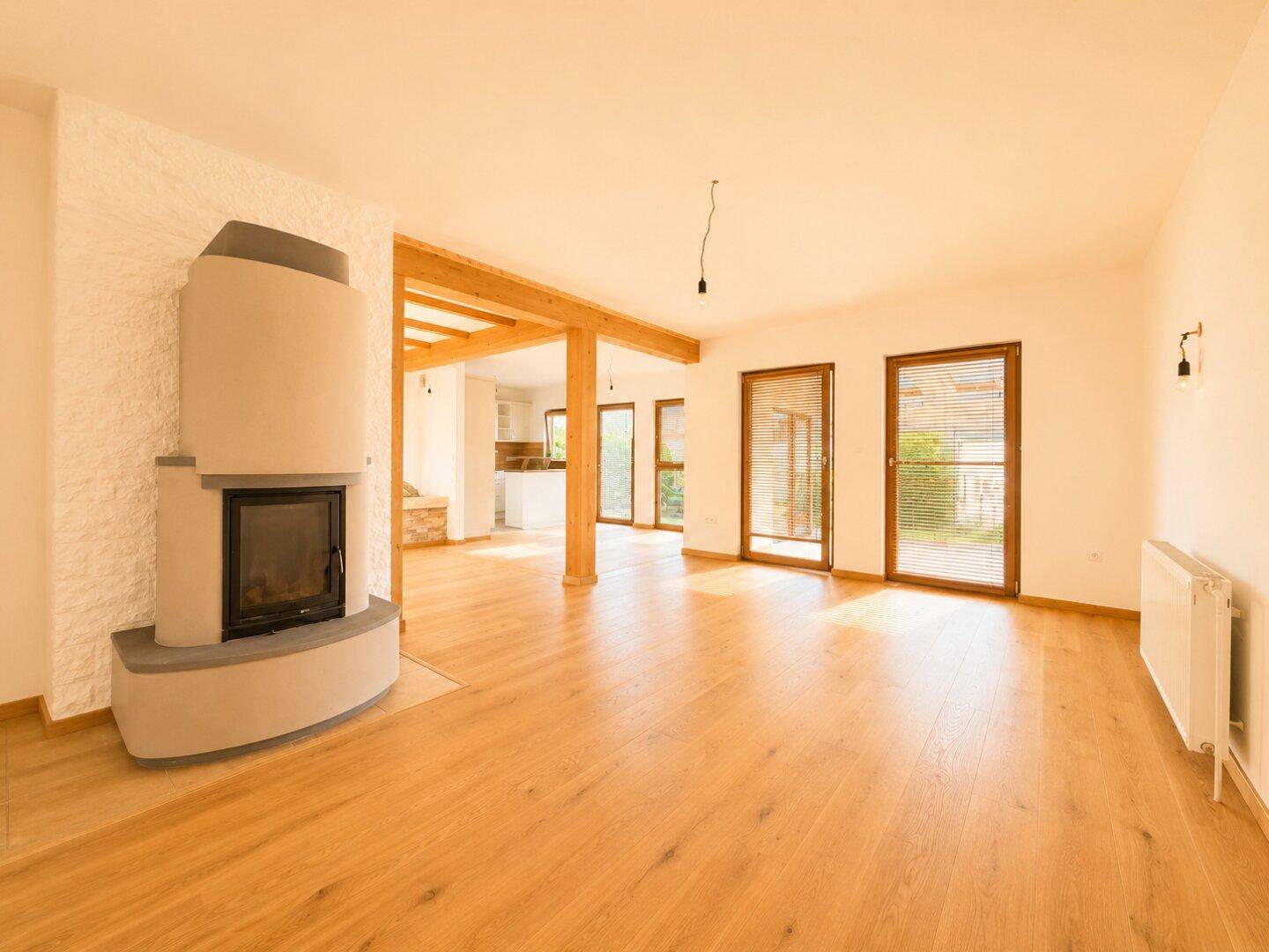 Heller Wohnbereich mit Kamin, Holzbalken und großen Fenstern mit Blick ins Grüne.
