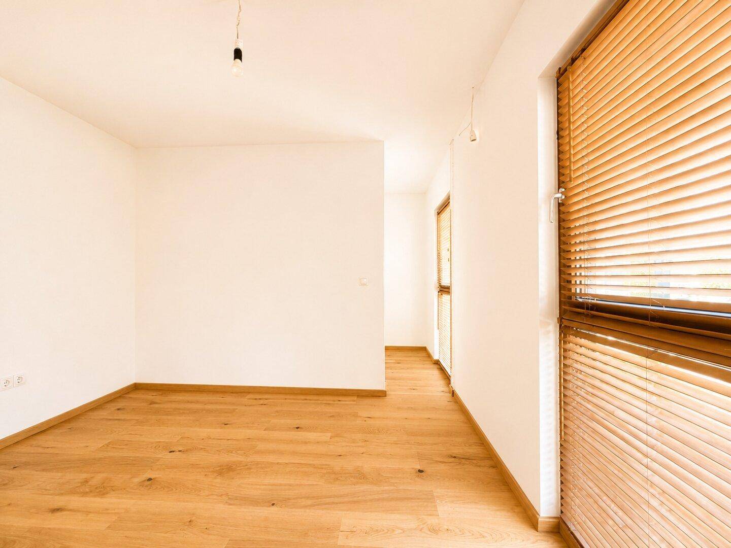 Helles, leeres Zimmer mit Eichenparkettboden und großen Fenstern mit Jalousien.