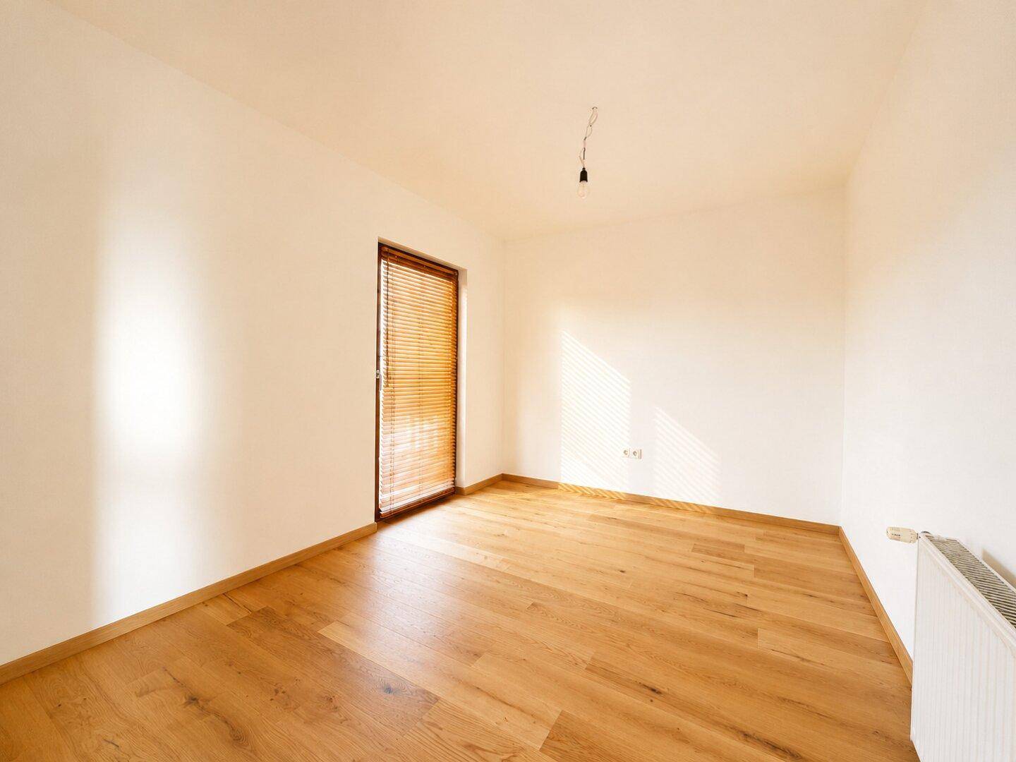 Sonniger Raum mit Holzboden und einem Fenster mit Jalousie, ideal für ein Schlafzimmer oder Büro.