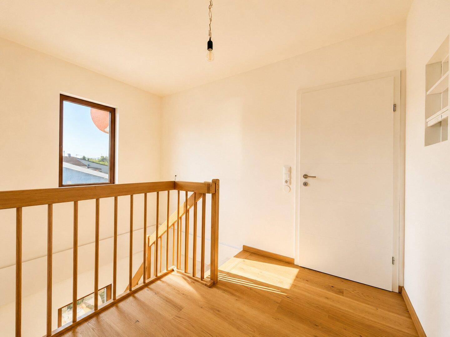 Heller Flur im Obergeschoss mit Holzgeländer und Blick nach draußen durch ein kleines Fenster.