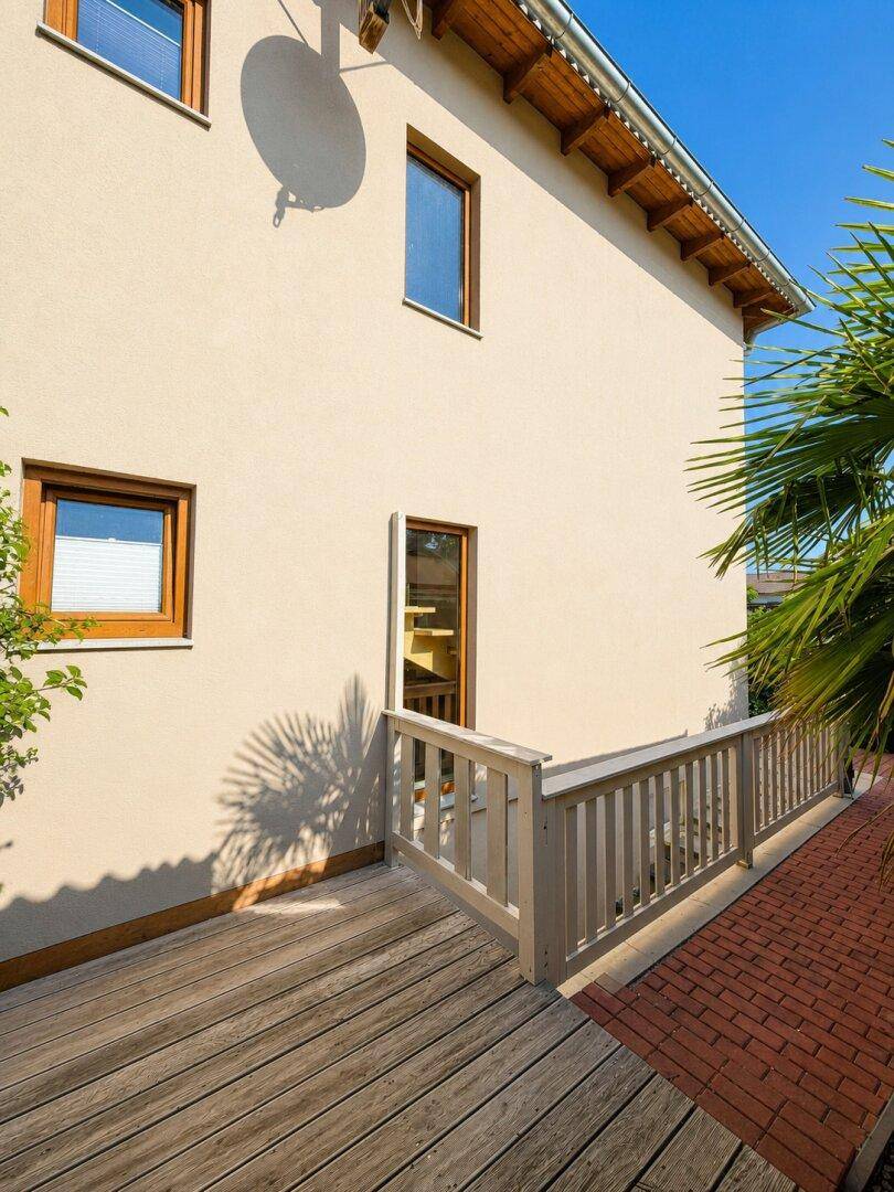 Seitlicher Zugang zum Haus mit Holzterrasse und Ziegelweg, umgeben von Palmen.