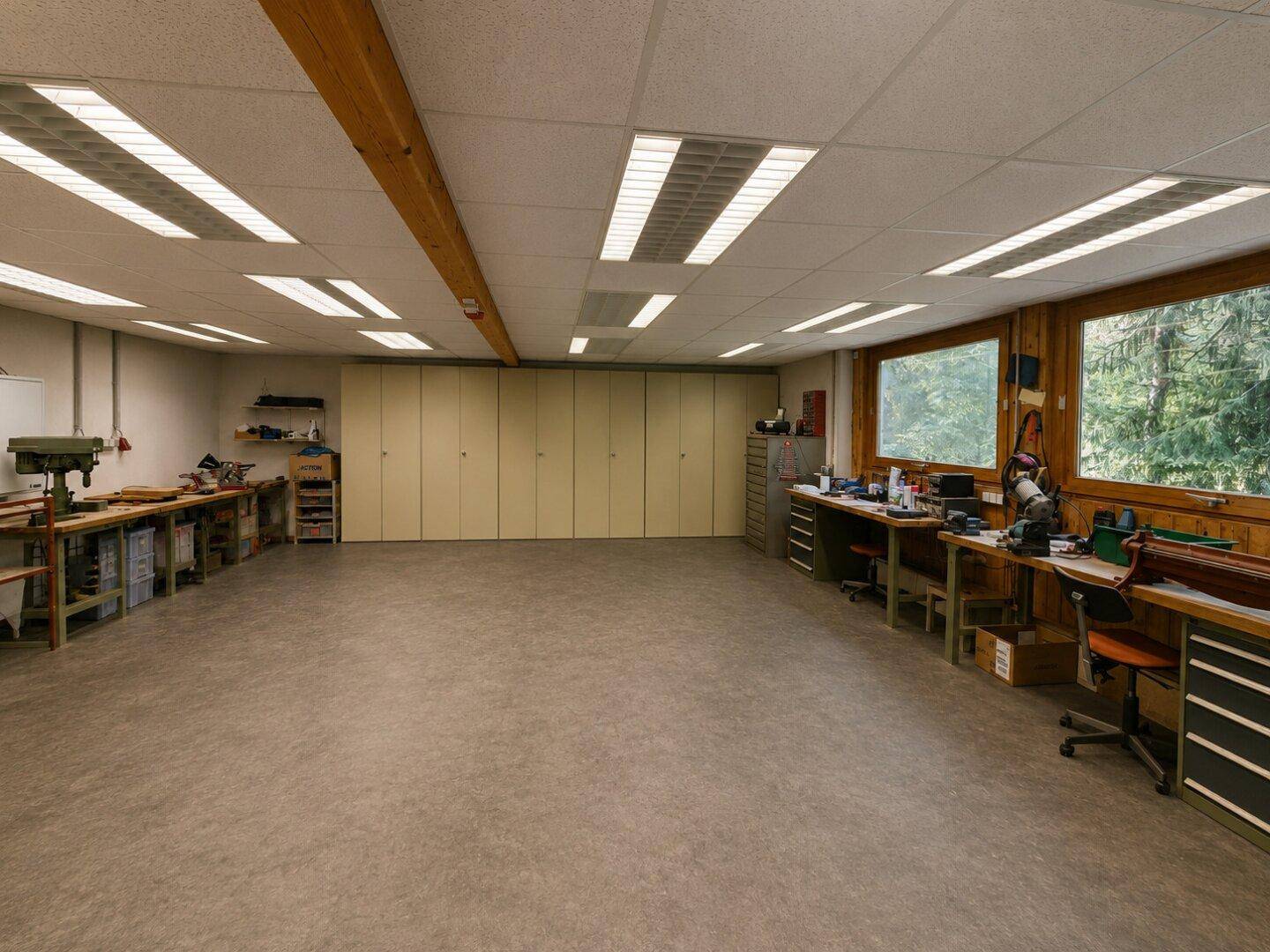Großer Werkstattraum mit Arbeitsbänken, Schränken und großen Fenstern mit Waldblick.