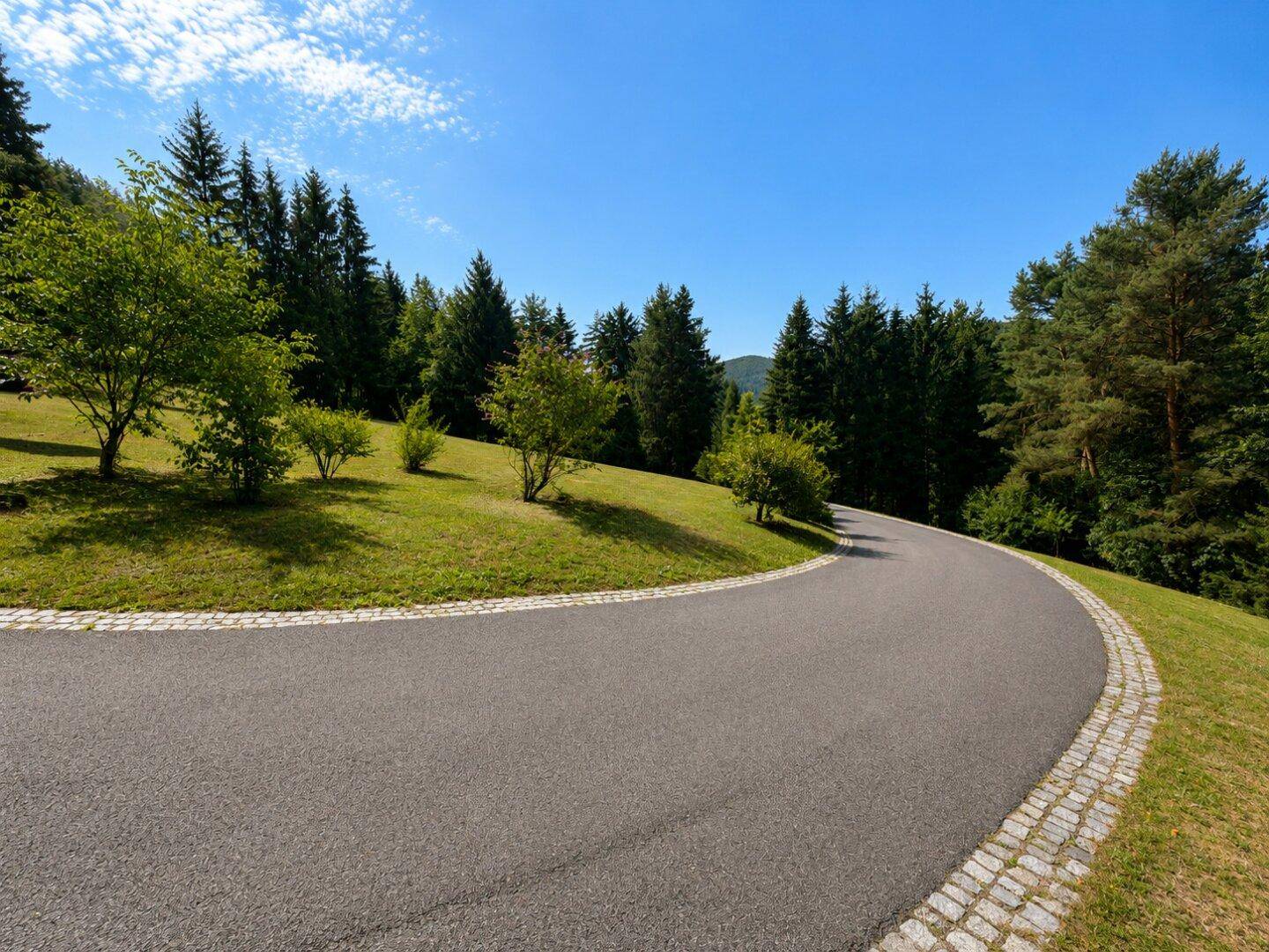 Geschwungene Asphaltstraße mit Kopfsteinpflasterrand, umgeben von grünen Wiesen und Bäumen.