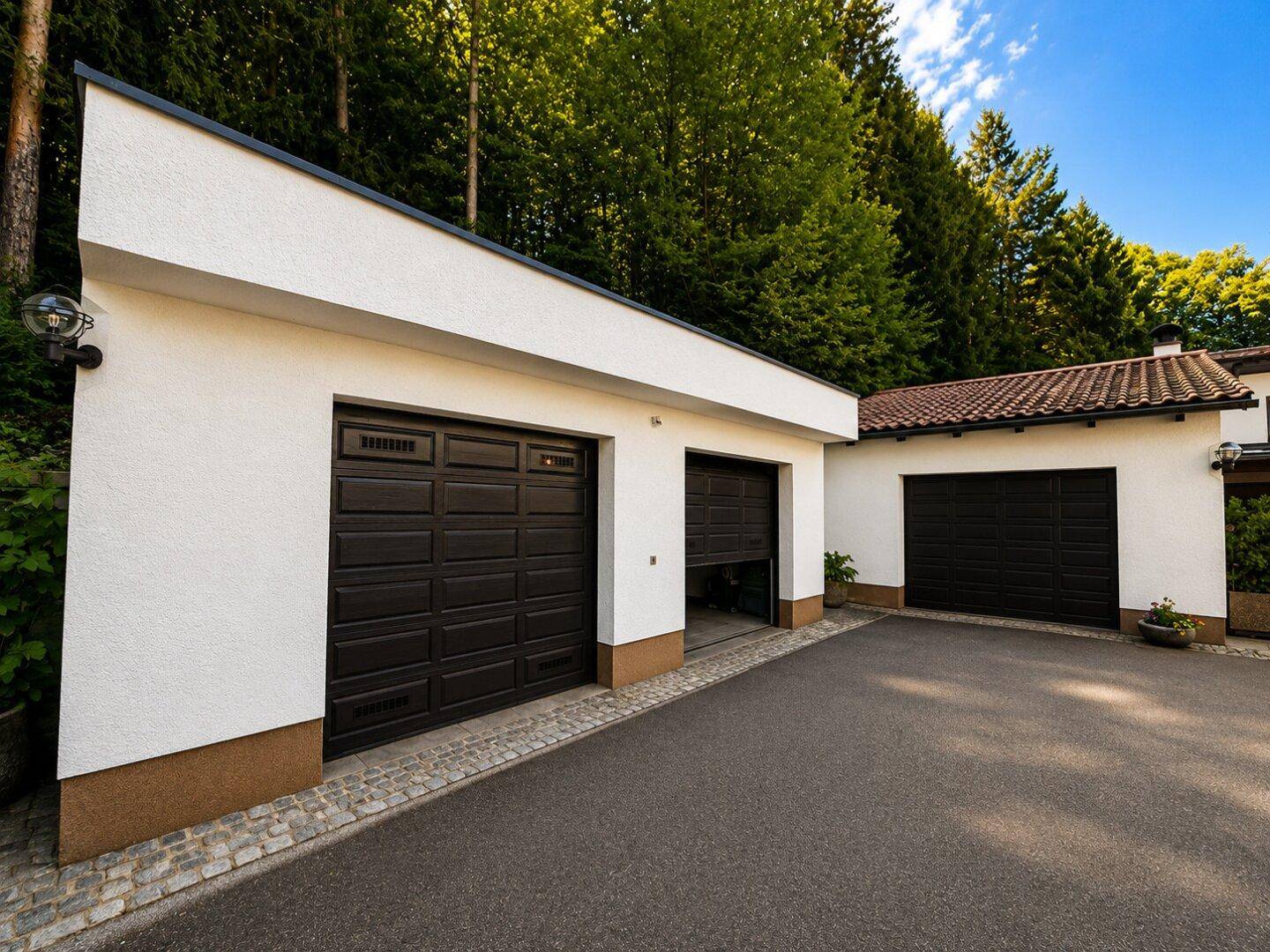 Zwei Garagen mit dunklen Toren und weißer Fassade, Teil des Anwesens mit Zufahrt.