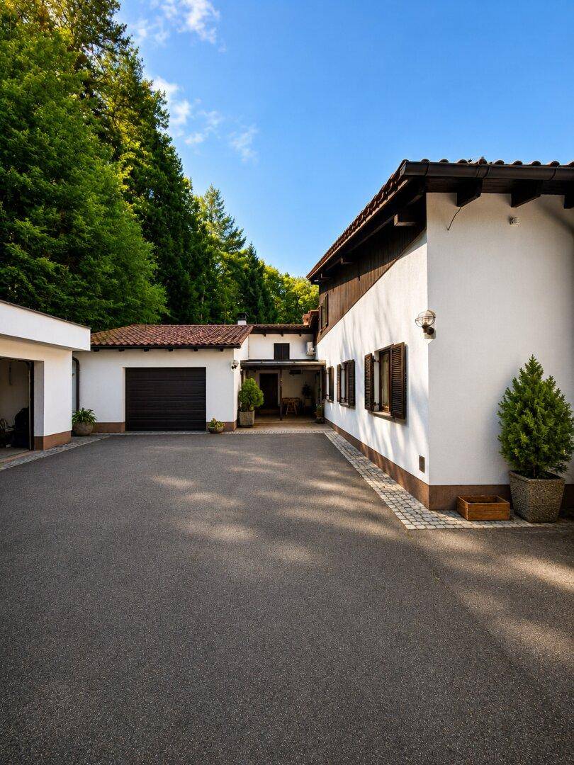 Ansicht des Hauses mit Zufahrt und Garage, eingebettet in eine grüne Umgebung mit hohen Bäumen.