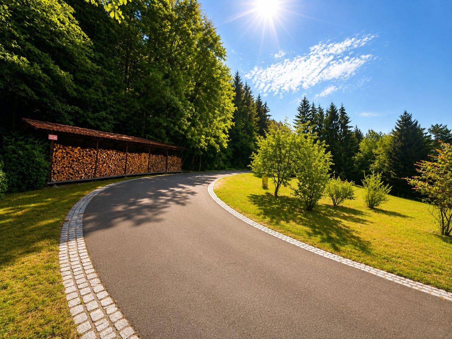 Zufahrt mit großem Holzschuppen, umgeben von Bäumen und grünen Hügeln unter blauem Himmel.