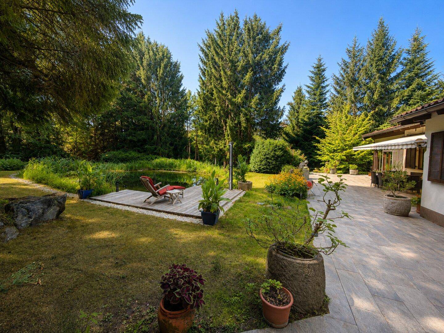 Weitläufiger Gartenbereich mit Teich, Holzdeck, Liegestuhl und angrenzender gefliester Terrasse am Haus.