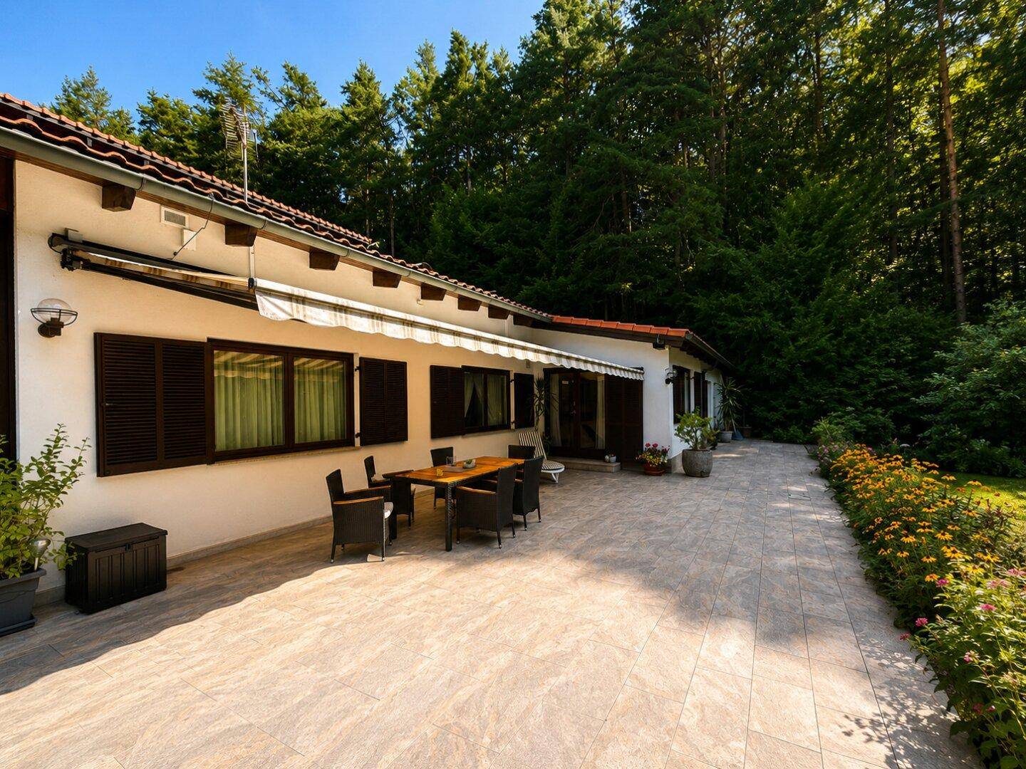 Große geflieste Terrasse mit Esstisch und Stühlen, umgeben von einem gepflegten Garten und hohen Bäumen.