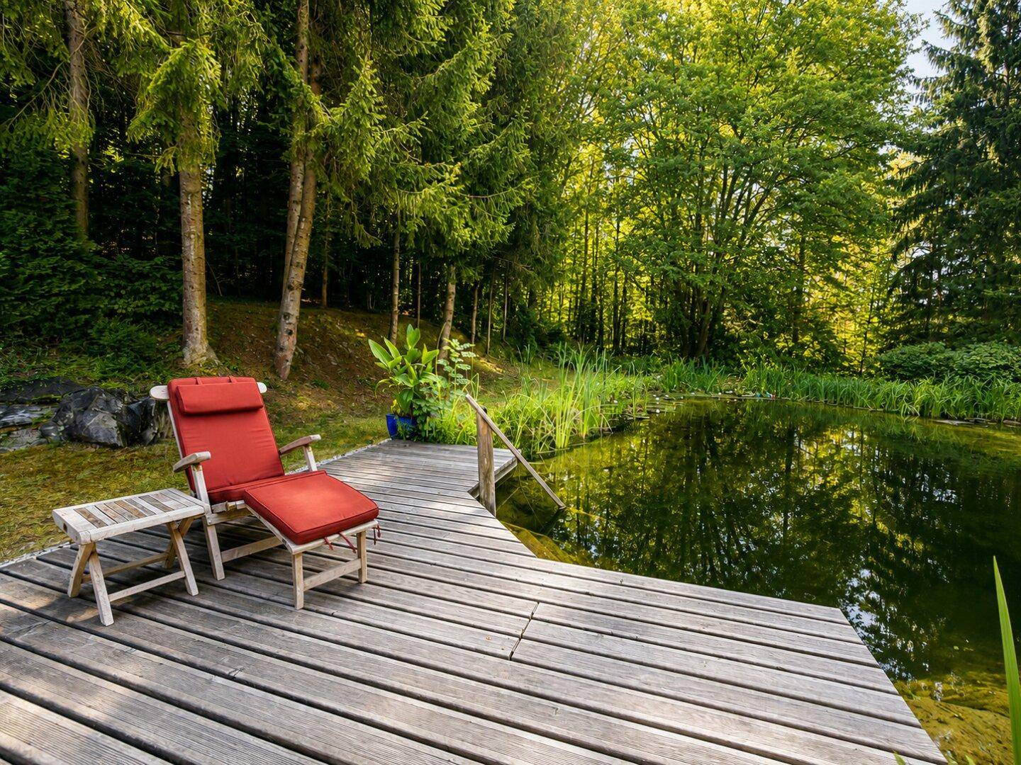 Holzdeck am Teich mit rotem Liegestuhl, umgeben von Bäumen und grünen Pflanzen.