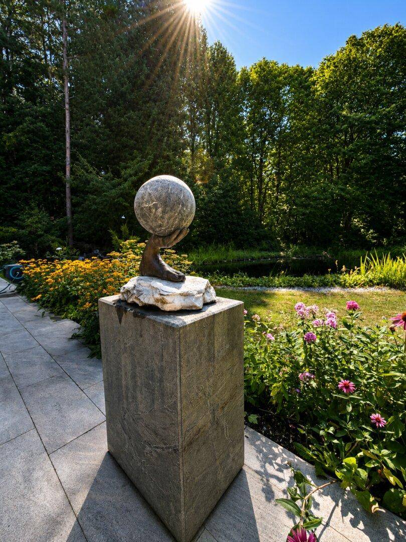 Sonnige Gartenterrasse mit Skulptur, Blumenbeeten und üppiger grüner Vegetation.