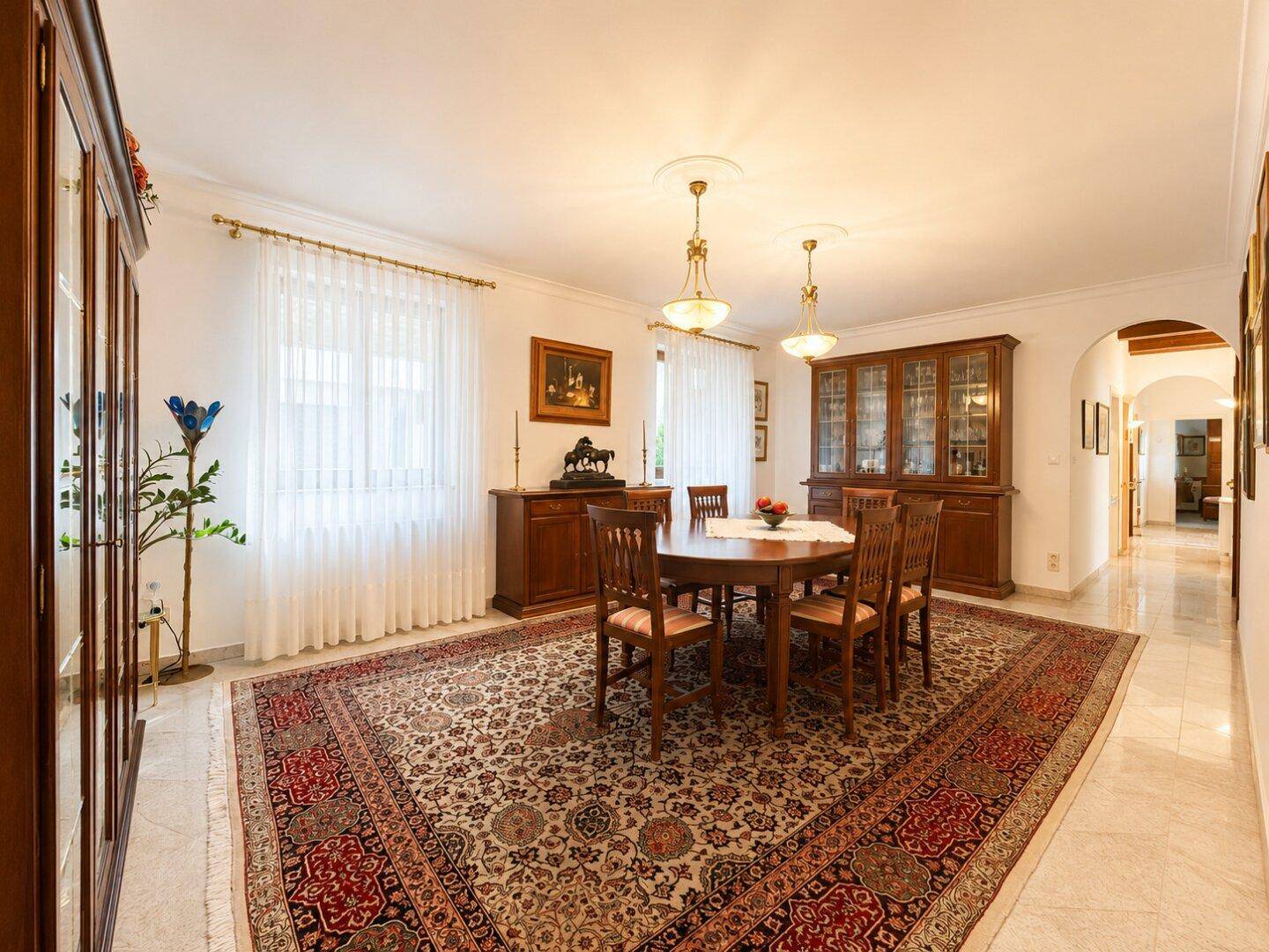 Esszimmer mit rundem Holztisch, traditionellen Stühlen, großem Teppich und einem Vitrinenschrank.