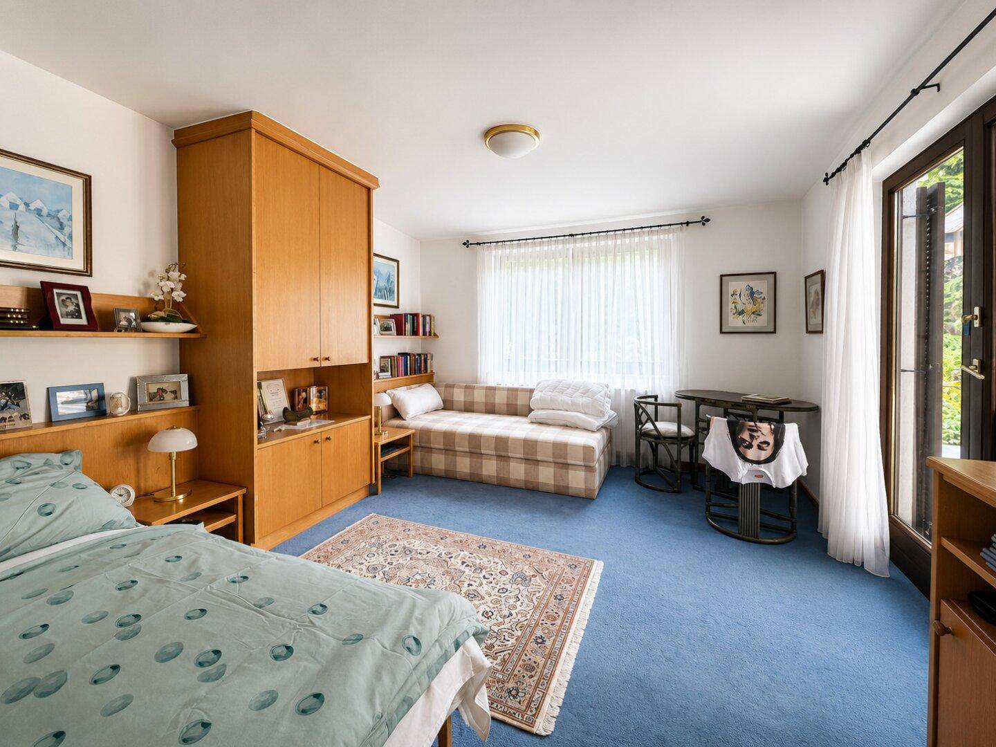 Schlafzimmer mit blauem Teppichboden, Holzschrank und Zugang zu einem Balkon oder Garten.